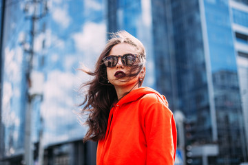 Closeup portrait of effective girl with curly hair on street on building background. brunette hair waving and shine on the sun rayin.bright orange sweatshirt and trendy sunglasses