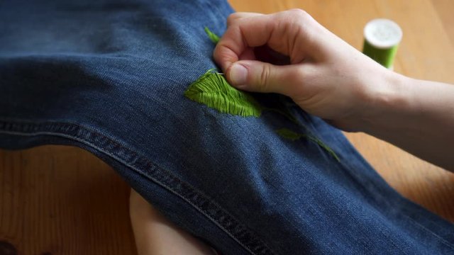 Close-up female hands making handmade color embroidery pattern on jeans using thread and sewing needle high angle. Art woman enjoying decorating design casual daily clothes