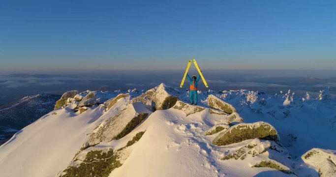 skier stands on top of the mountain, lifting skis up and shaking them. above the clouds. the camera's flying at him. aerial view drone. 