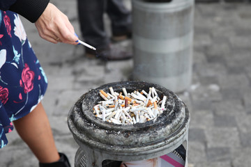 Cigarette in the hands of a woman near an ashtray with lots of cigarette butts