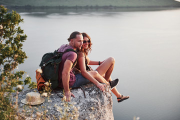 Hugging couple with backpack sitting on top of rock mountain enjoying view coast a river or lake. Traveling along mountains and coast, freedom and active lifestyle concept