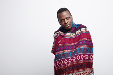 Authentic portrait of young handsome African American guy, kindly looking at camera, wearing interesting ethnic poncho, smiling gently, isolated over white background with copy space for advertising.