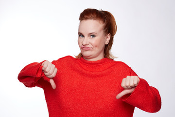 Red haired female expresses dislike with body language, keeps thumbs down and frowns face, has displeased face, freckled skin, curves lips, isolated over white background. This is really bad idea!