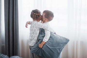 Happy kids playing in white bedroom. Little boy and girl, brother and sister play on the bed wearing pajamas. Nightwear and bedding for baby and toddler. Family at home