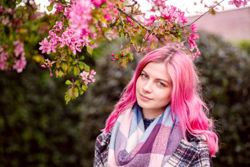 Fototapeta premium young beautiful girl with pink hair standing near a tree with flowers, pink flowers, spring, sun, happiness