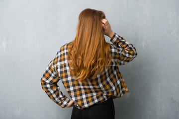 Young redhead girl over grunge wall on back position looking back while scratching head