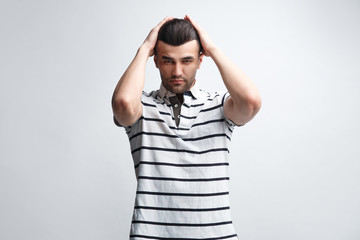 An attractive man in a striped polo shirt holds his head in his hands against a light gray...