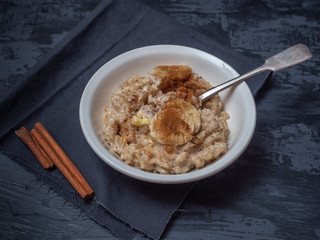 A plate of porridge with a sprinkling of cinnamon and banana circles. A teaspoon in a plate with each, a black cotton napkin. Nearby are cinnamon sticks.