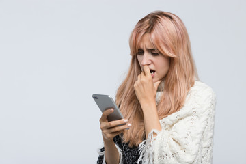 Studio portrait of blonde teen in white sweater waiting for a response sms, gnawing nails, looking...