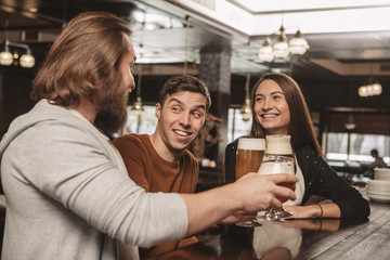 Handsome young man smiling happily, enjoying resting at the bar with his friends. Group of friends having drinks at the local beer pub. Two men and beautiful woman drinking beer. Celebration concept