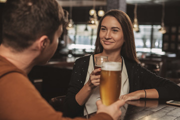 Beautiful happy woman smiling cheerfully, talking to her boyfriend over a glass of beer at the local pub. Lovely young female drinking delicious craft beer with her friend at the bar. Dating concept