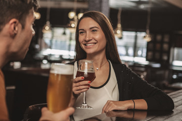 Gorgeous brunette young woman laughing, enjoying talking to her man, relaxing at the bar together, having drinks. Happy couple enjoying drinking craft beer at the pub. Love, flirt, romance concept