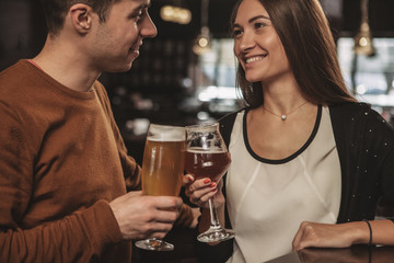 Cropped shot of a happy loving couple clinking their beer glasses, smiling at each other. Cheerful man and his beautiful girlfriend having drinks at the bar, celebrating Valentines Day. Love, couples 