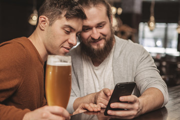 Two male friends enjoying resting at beer pub together, checking out news online, using smart phone. Bearded man browsing on the internet, showing something to his friend. Social media, online concept