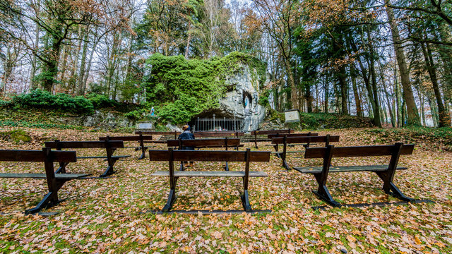 image of a woman sitting on a bench outdoors praying to the Virgin of Lourdes that is in a natural grotto with trees in the background on a magical autumn day in Vielsalm in the Belgian Ardennes