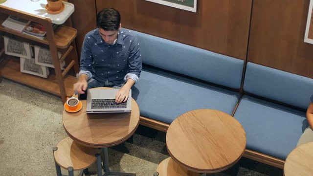 Attractive hispanic man sitting in coffee shop or hotel lobby with laptop and cell phone drinking coffee, top view