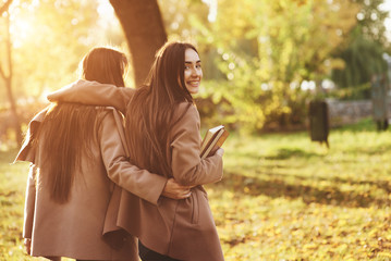 View from the back of young smiling brunette twin girls hugging and having fun in casual coat walking at autumn sunny park on blurry background. One of them is looking backwards and holding books - obrazy, fototapety, plakaty