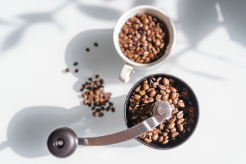 A white cup filled with coffee beans next to a coffee grinder and loose single beans on a bright glass table in a drop view. the sunlight comes from the side