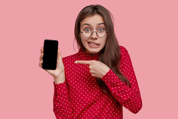 Naklejka premium Puzzled dark haired young woman in optical glasses, points at electronic gadget with mock up screen, wears red shirt, advertises new device, has green eyes, models over pink background, uses app