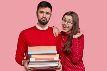 Dissatisfied man with upset look, carries pile of books, tired of studying, happy Caucasian girlfriend expresses good emotions, stand closely, isolated over pink background. Learning concept
