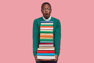 Emotional surprised black man looks with terrified expression, carries pile of textbooks, afraids...