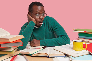 Horizontal shot of surprised dark skinned man wears optical glasses, focused with frightened expression at opened textbook, learns new material, isolated over pink wall. Student prepares for seminar