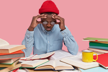 Concentrated puzzled dark skinned man in red headgear, optical round glasses, elegant shirt, focused with worried expression at textbooks, busy working and doing home assignment, isolated on pink
