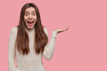 Studio shot of happy dark haired woman raises palm, feels excited, holds copy space, dressed casually, demonstrates something, satisfied to show something awesome and cool, isolated on pink wall