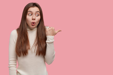 Portrait of attractive young woman points with thumb at blank copy space, stares at camera, has bated breath, dressed casually, models over pink background, empty space for your promotional content