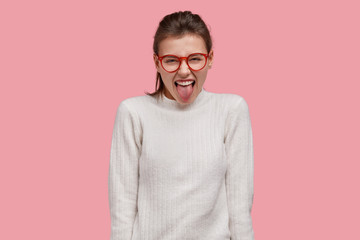 Studio shot of attractive young woman shows tongue, makes grimace, foolishes indoor, wears...