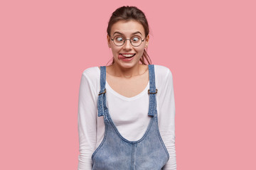 Fototapeta premium Photo of happy woman crosses eyes, shows tongue, foolishes with friend, dressed in fashionable clothes, stands against pink background. Photo of attractive girlfriend makes grimace. Facial expressions