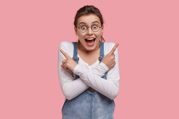 Glad Caucasian woman with joyful expression, points at different sides, wears casual clothes, hesitates what to choose, isolated over pink background, feels uncertain between choices. Which way?