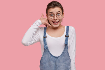 Happy young woman makes call gesture, has satisfied expression, keeps hand near ear, looks through round spectacles, dressed in jean sundress, models over pink background. Call me back, please