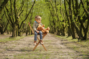Naklejka na ściany i meble A lovely, smiling boy sitting on the chair with teddy bear at the springtime. Hazel tree