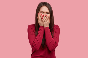 Depressed brunette lady covers mouth with hands, has mournful expression, black hair, dressed in red clothes, poses against pink background, regrets something. Grieved lady cries with depression