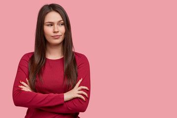 Fototapeta premium Photo of thoughtful brunette young woman with dark hair, keeps arms folded, considers something in mind, wears red sweater, stands against pink background. People, thoughts, considering concept
