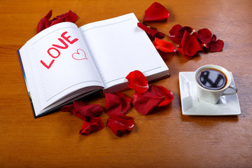 A cup of coffee and a diary with the word love written in red on paper on a wooden table covered with rose petals.