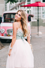 View from back pin up styled girl with long blonde hair walking on street. She wears light tulle skirt, has bare shoulder, smiling to camera