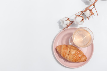 A branch of cotton on a light background and a croissant on a pink plate. Top view. Copy space.