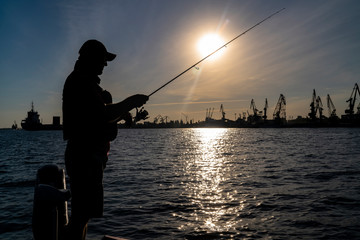Urban fishing concept. Silhouette of fisherman on industrial seaport city background.