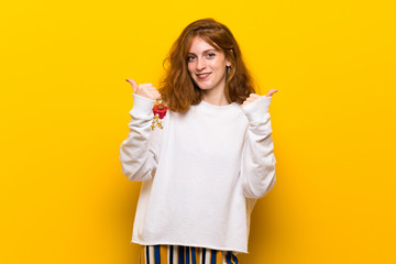 Young redhead woman over yellow wall giving a thumbs up gesture with both hands and smiling