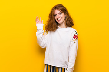 Young redhead woman over yellow wall saluting with hand with happy expression