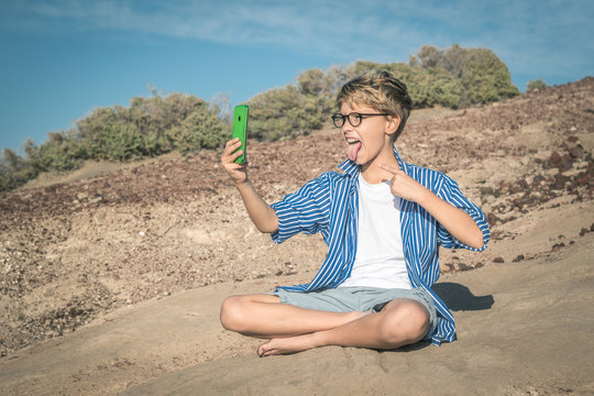 Blond child with eyeglasses on the beach takes a selfie, wearing striped shirt and sitting cross-legged. Taking picture of bigmouth with cell. smartphone generation. Blue sky and rocks in background
