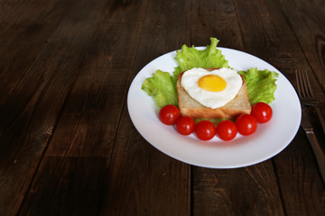 fried eggs with a yellow yolk in the form of a heart on bread, decorated with green salad and red cherry tomatoes on a white plate and wooden rustic background with space for text 