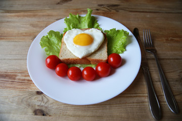 fried eggs with a yellow yolk in the form of a heart on bread, decorated with green salad and red cherry tomatoes on a white plate and wooden rustic background 