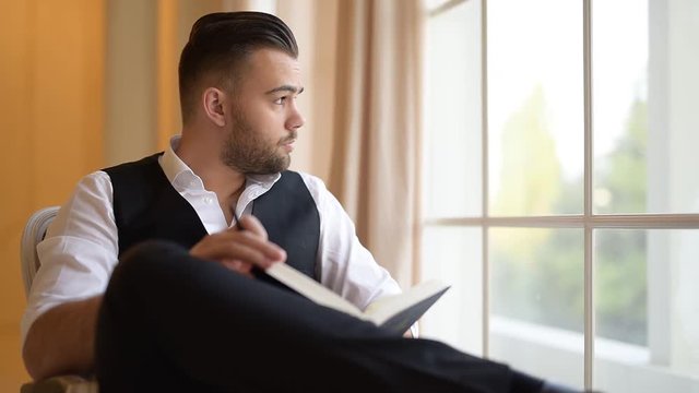 A young man sitting on armchair and flips through a book ove background of window.