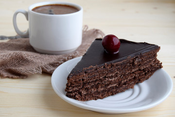 The piece of chocolate cake with cherry is on a white plate. The cup with coffee is near cake.