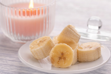 Sliced banana on a white plate and a light wooden table. Pink burning candle nearby.