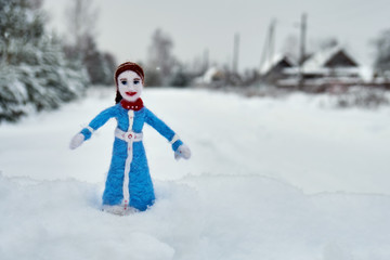 Needle felted wool doll stands on the snow with a russian village on the background