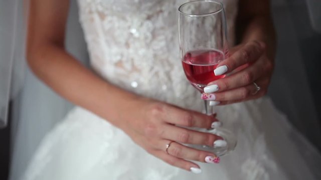 The bride in a white dress is holding a glass of pink champagne standing on a gray background. A beautiful thin glass of pink wine in the hands of a lovely sweet bride.